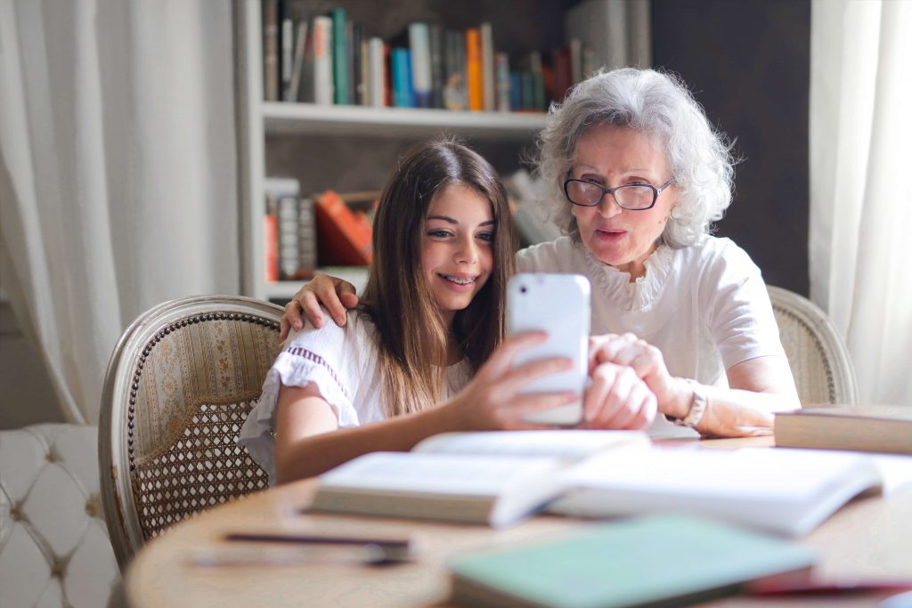 Seniorin und Enkelkind sitzen am Tisch und schauen auf Smartphone. Es soll die Technikaffinität von Senioren darstellen.