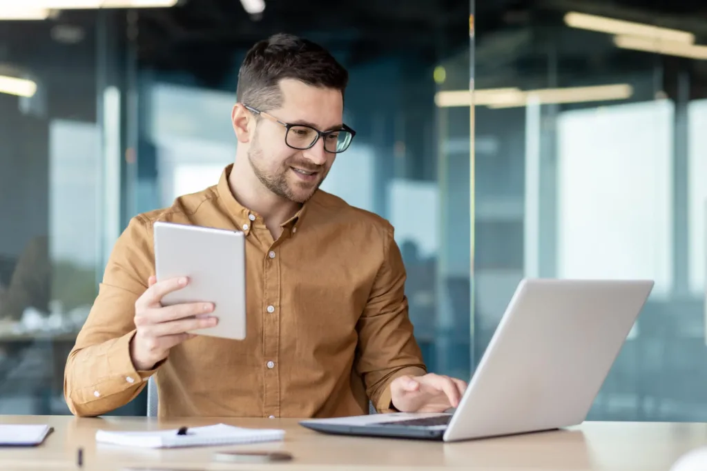 Ein Mann führt am Laptop ein Videotelefonat und hält zusätzlich ein Tablet in der Hand.
