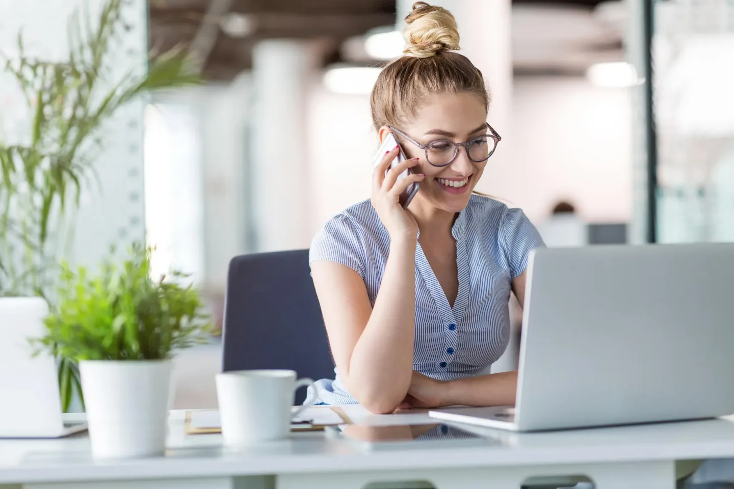 Symbolbild – besser telefonieren mit TeleSys – Junge Frau nutzt klassische Telefonanlage um störungsfrei zu telefonieren und lächelt dabei positiv in ein Laptop