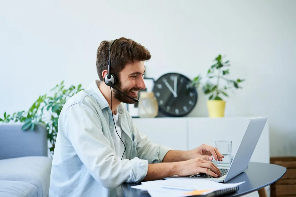 Mann arbeitet im Homeoffice im Wohnzimmer am Laptop und telefoniert mit einem Headset.